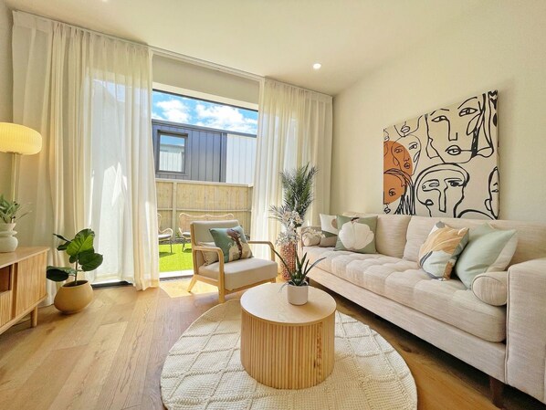 A modern living room with a sofa, coffee table, and a chair. There is a large window with curtains, a painting on the wall, and a potted plant.