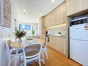 Une cuisine moderne avec des appareils électroménagers blancs, un coin repas avec des chaises en osier et une vue sur l’extérieur.