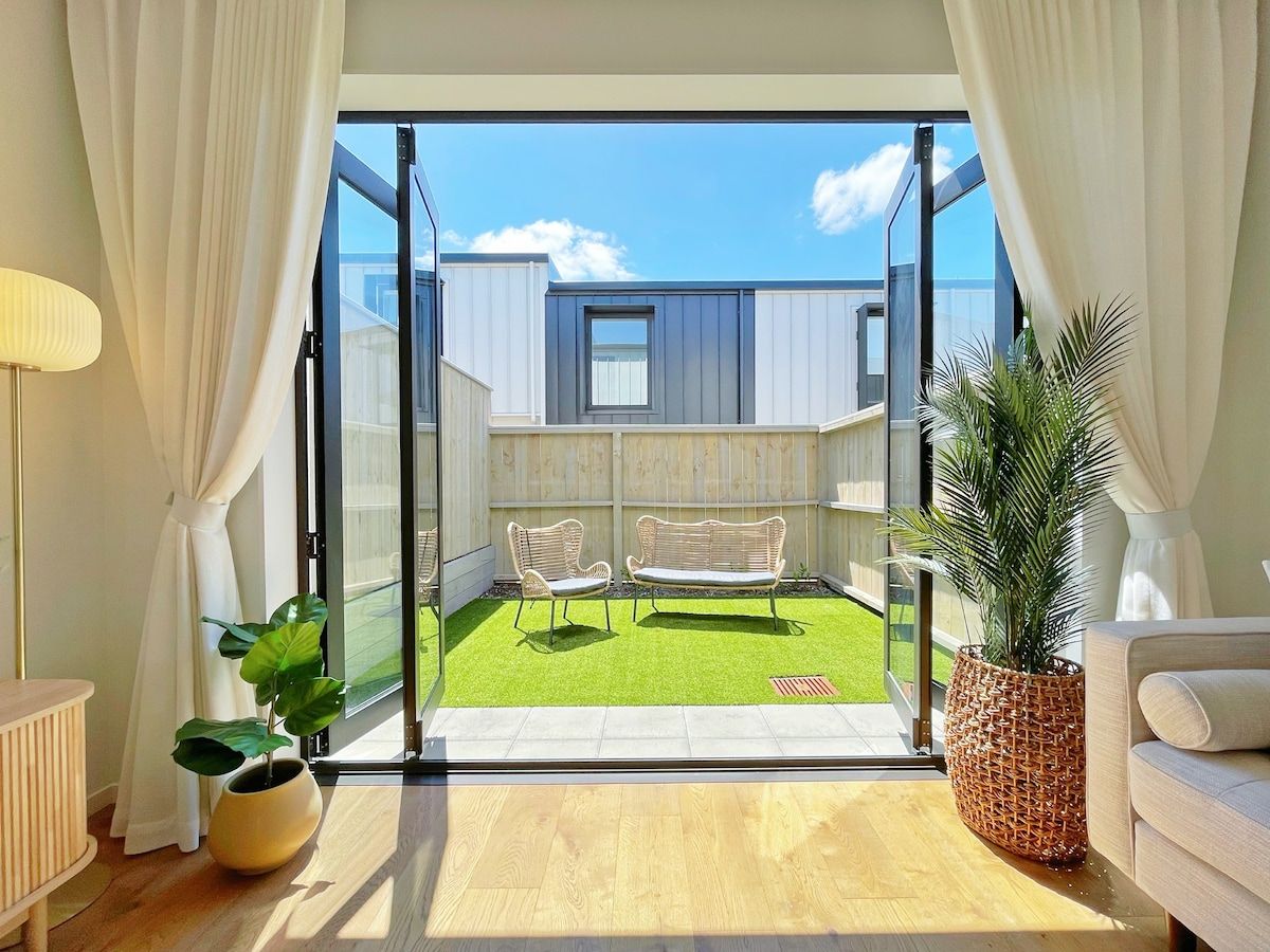 Un salón moderno con un ventanal corredero que da a un jardín con césped artificial y dos sillones de mimbre.
