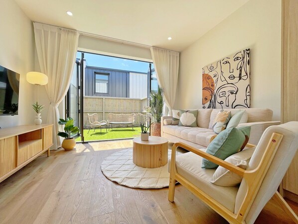 A modern living room with a sofa, armchair, and coffee table, a large window with curtains, and a view of a balcony.