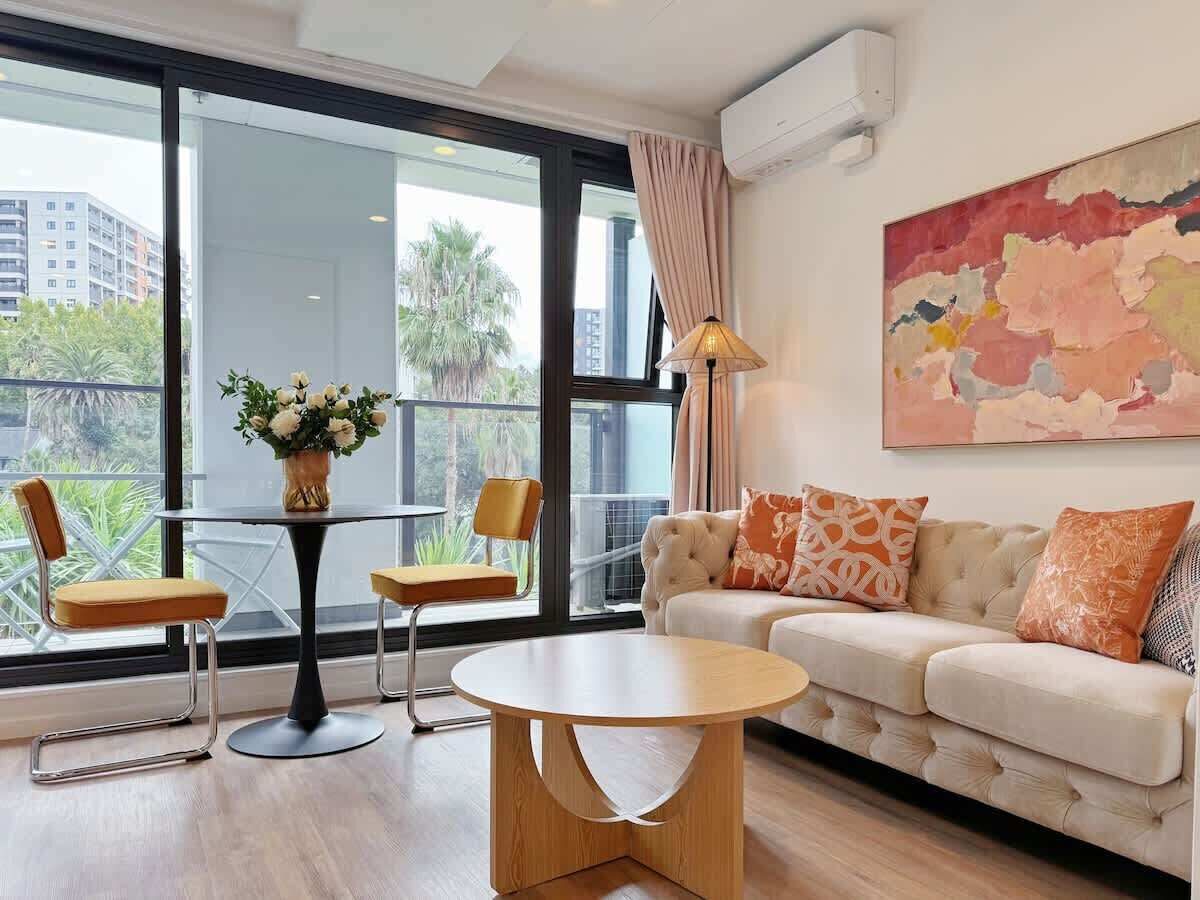 Una sala de estar moderna con un sofá, dos sillones, una mesa de centro y una mesa de comedor con sillas. Hay un ventanal con vistas a edificios y árboles.