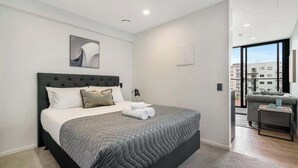 Un dormitorio moderno con una cama grande, cabecera, mesita de noche y vistas de la ciudad a través de un amplio ventanal.