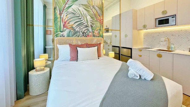 Hotellihuone, jossa on suuri sänky, trooppisten lehtien kuva, pieni keittiönurkkaus ja kylpyhuone.