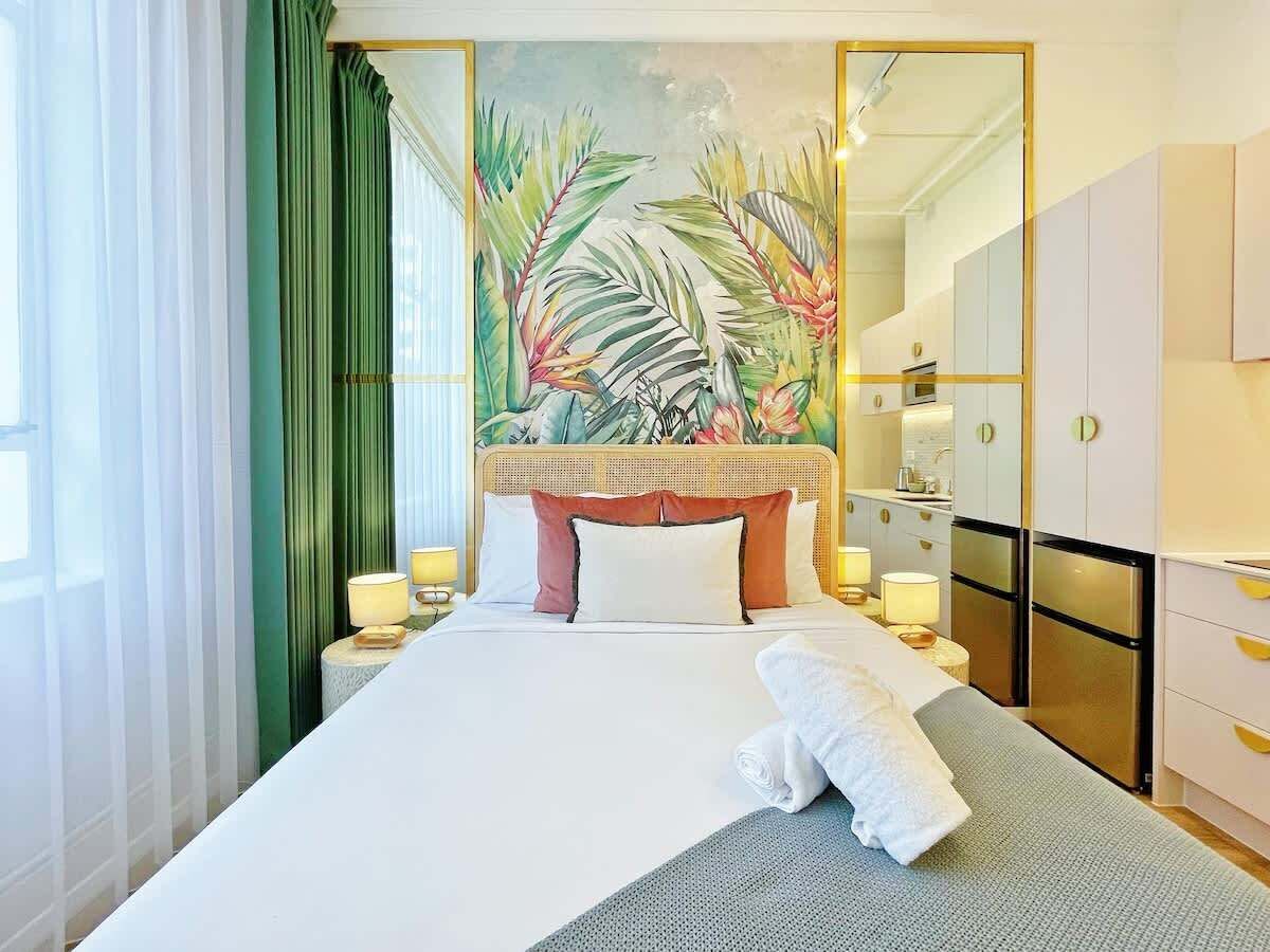 A hotel room with a large bed, a painting of tropical foliage on the wall, a wardrobe, and a nightstand with lamps.