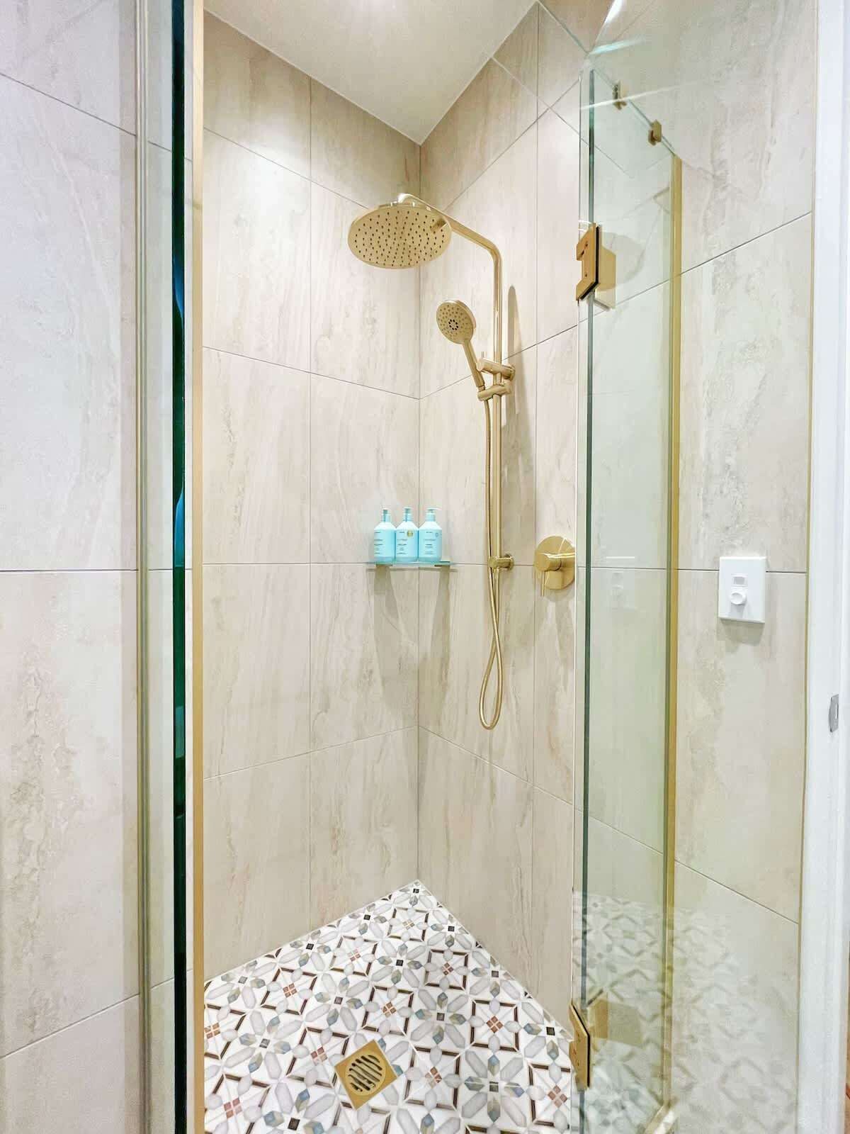 Une salle de bain moderne avec une douche à l’italienne en verre, une pomme de douche dorée et un carrelage décoratif.