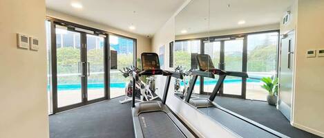 Une salle de sport moderne équipée de tapis de course et de vélos d’exercice, avec de grandes fenêtres donnant sur une piscine.