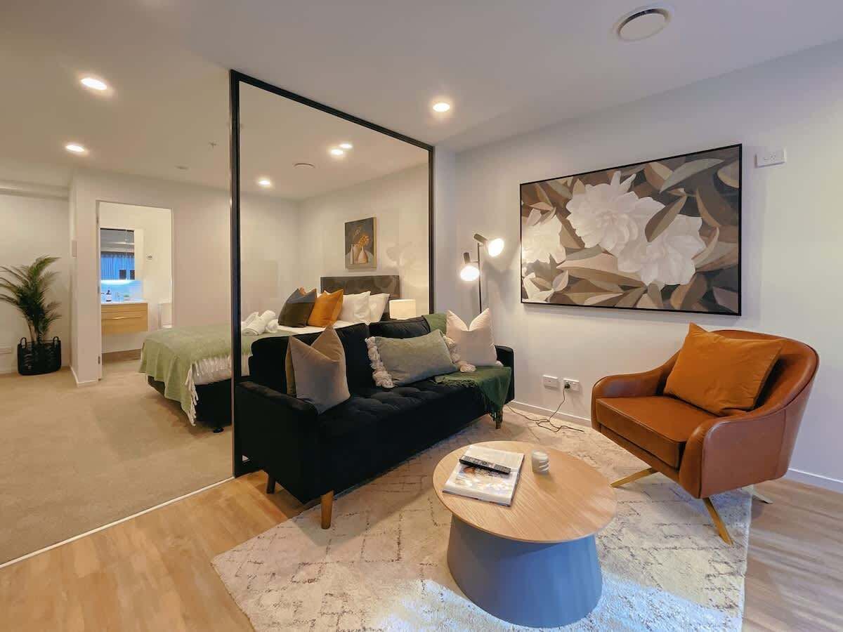 Un salon moderne avec un canapé noir, un fauteuil orange et une table basse ronde. Un grand miroir reflète la pièce, et un tableau est accroché au mur.