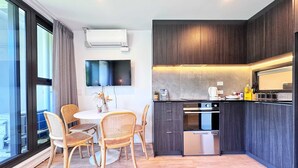 Una cocina moderna con armarios de madera oscura, horno empotrado y lavavajillas de acero inoxidable. Hay un comedor con sillas de mimbre y una mesa redonda, y un televisor de pantalla plana montado en la pared.