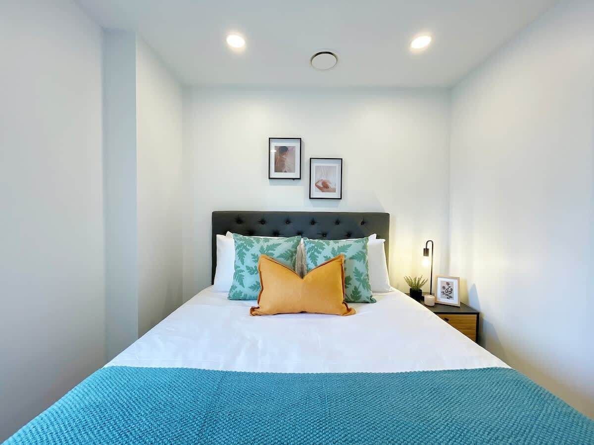 Un lit bien fait, recouvert d’une couvre-lit blanche et bleue, avec deux oreillers et une tête de lit. Deux tableaux encadrés sont accrochés au mur, au-dessus du lit.