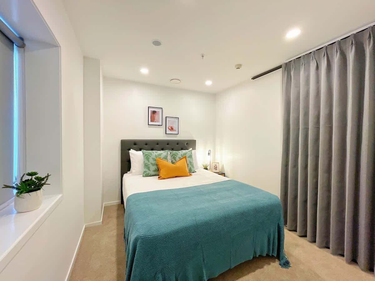 Un dormitorio moderno con cama, cabecera, ventana con persianas, cortina y una pequeña planta en maceta.