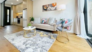 A modern living room with a sofa, a coffee table, and a chair. There is a kitchen in the background with a window. - City & Ponsonby Center Side 2 BDR Townhouse vs AC (Auckland)