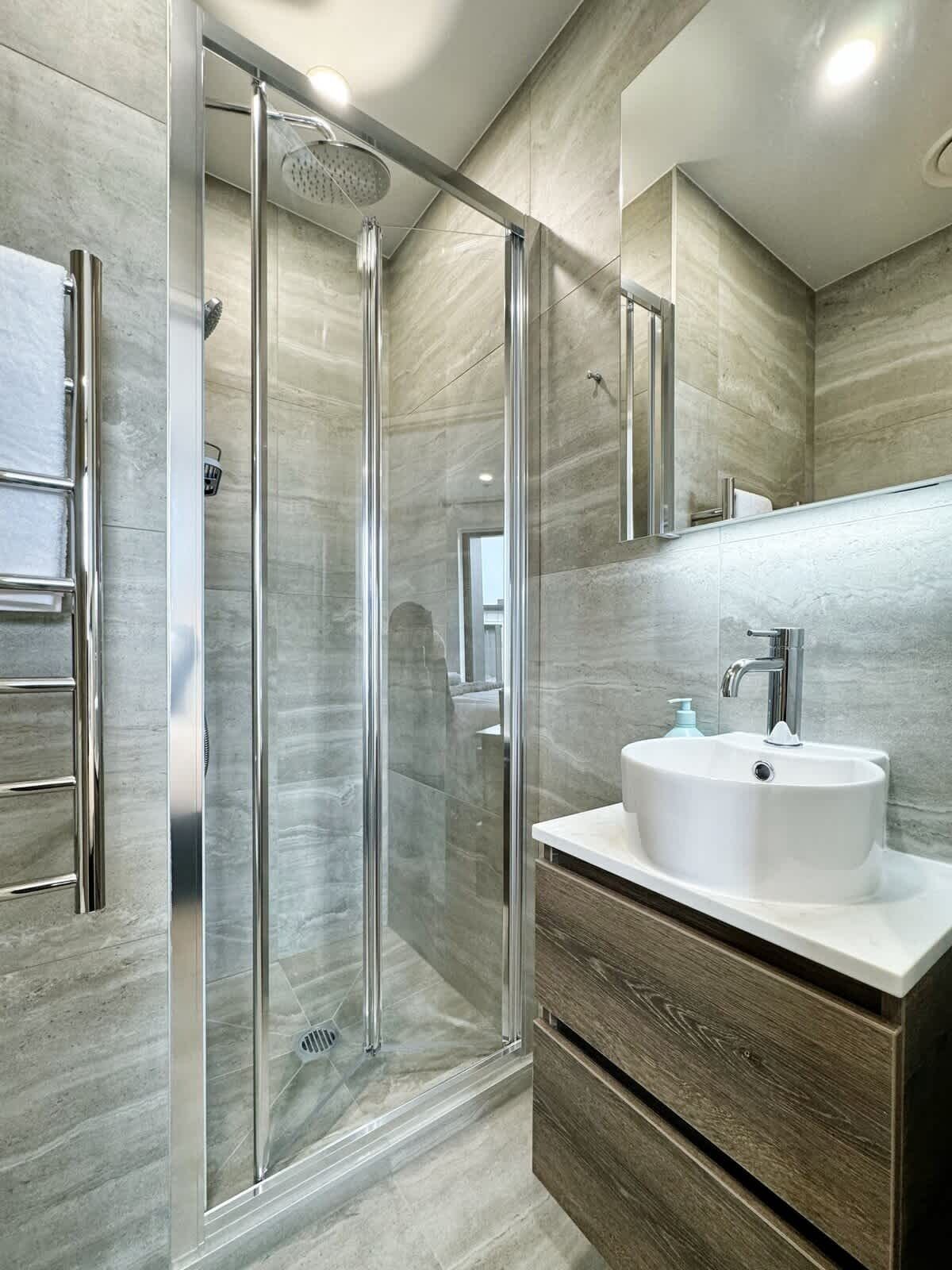 Un baño moderno con ducha envuelta en vidrio, un lavamanos blanco y un mueble de tocador de madera.