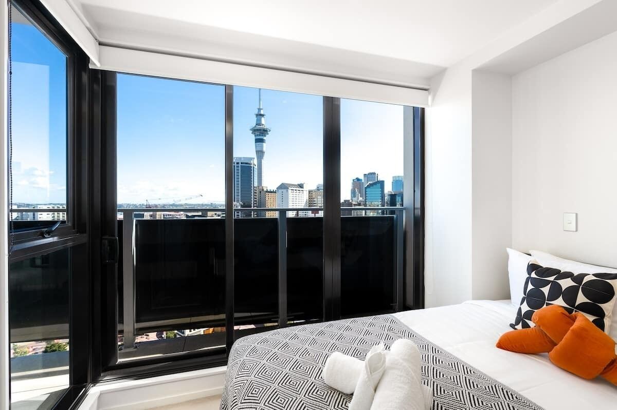 Une chambre d’hôtel moderne dotée d’une grande fenêtre offrant une vue panoramique sur la ville, d’un lit soigneusement fait avec une literie à motifs, et d’un paysage urbain visible à travers la fenêtre.