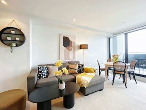 A modern living room with a grey sofa, black coffee table, and yellow flowers. There is a dining area with a table and chairs. - Incredible City & Skytower View Near Queen St-Pool (Auckland)