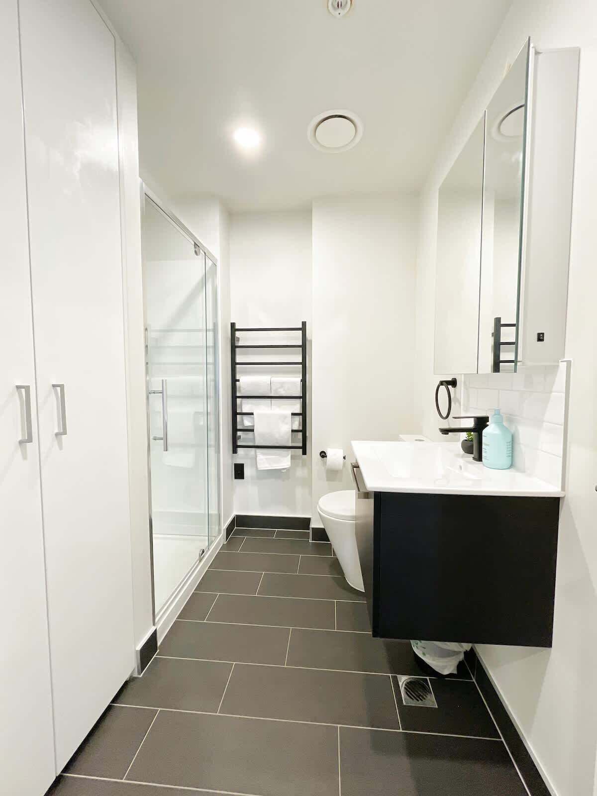 A modern bathroom with a glass shower enclosure, a black towel rack, a white toilet, and a black sink with a mirror above it.