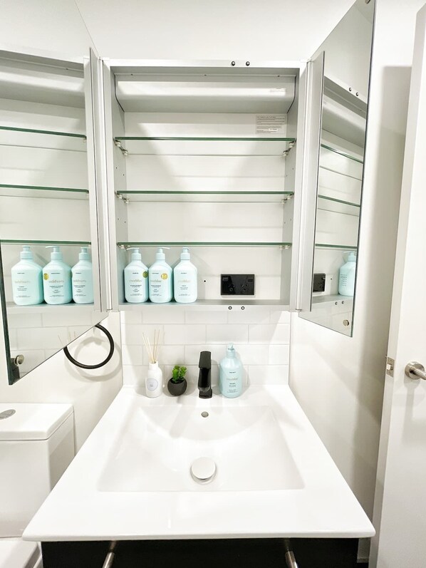 Un baño moderno con un lavamanos blanco, armarios con espejo y estantes con artículos de aseo.