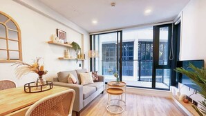 A modern living room with a sofa, dining table, and chairs. There are shelves with decorative items and a large window with a view.