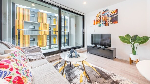 A modern living room with a sofa, a coffee table, a TV, and a large window overlooking a building with balconies. - Central Stylish Comfy Studio with Swimming Pool (Auckland)
