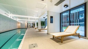 Ein moderner Poolbereich im Innenbereich mit einer großen Glaswand, Holzliegen und einem gefliesten Boden.