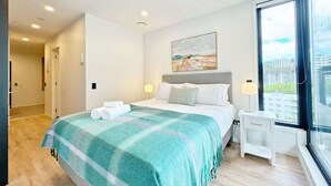 A modern bedroom with a large bed, a nightstand, a desk, and a window with a city view. - Lush City Escape - 1B Studio Near Skytower vs Pool (Auckland)