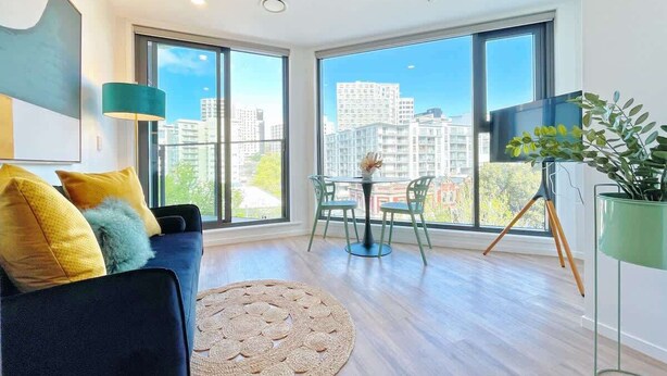 A modern living room with a sofa, a round rug, and a dining area with chairs and a table. - Parkside Elegance 1BR on Queen St vs Pool & Gym (Auckland)