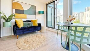 A modern living room with a dark blue sofa, yellow and gray cushions, a woven rug, a green chair, and a table with a vase of flowers. - Parkside Elegance 1BR on Queen St vs Pool & Gym (Auckland)