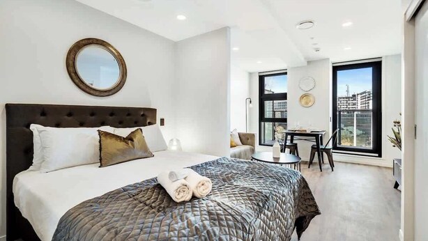A modern bedroom with a large bed, a round mirror, a dining area with a table and chairs, and a large window offering a city view. - Beautiful Designed Apartment with Pool Gym Sauna (Auckland)