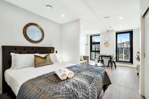 A modern bedroom with a large bed, a round mirror, a dining area with a table and chairs, and a large window offering a city view.