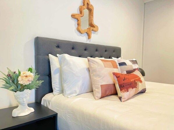 A neatly made bed with a grey headboard, white bedding, and a variety of decorative pillows. A white vase with flowers is placed on a bedside table.