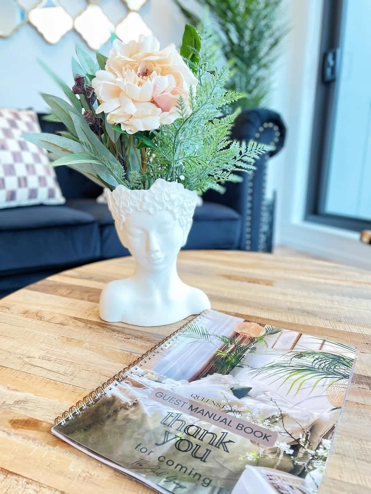 Eine weiße Vase mit Blumen auf einem Holztisch, ein Gästebuch und ein kariertes Kissen auf einer dunklen Couch im Hintergrund.