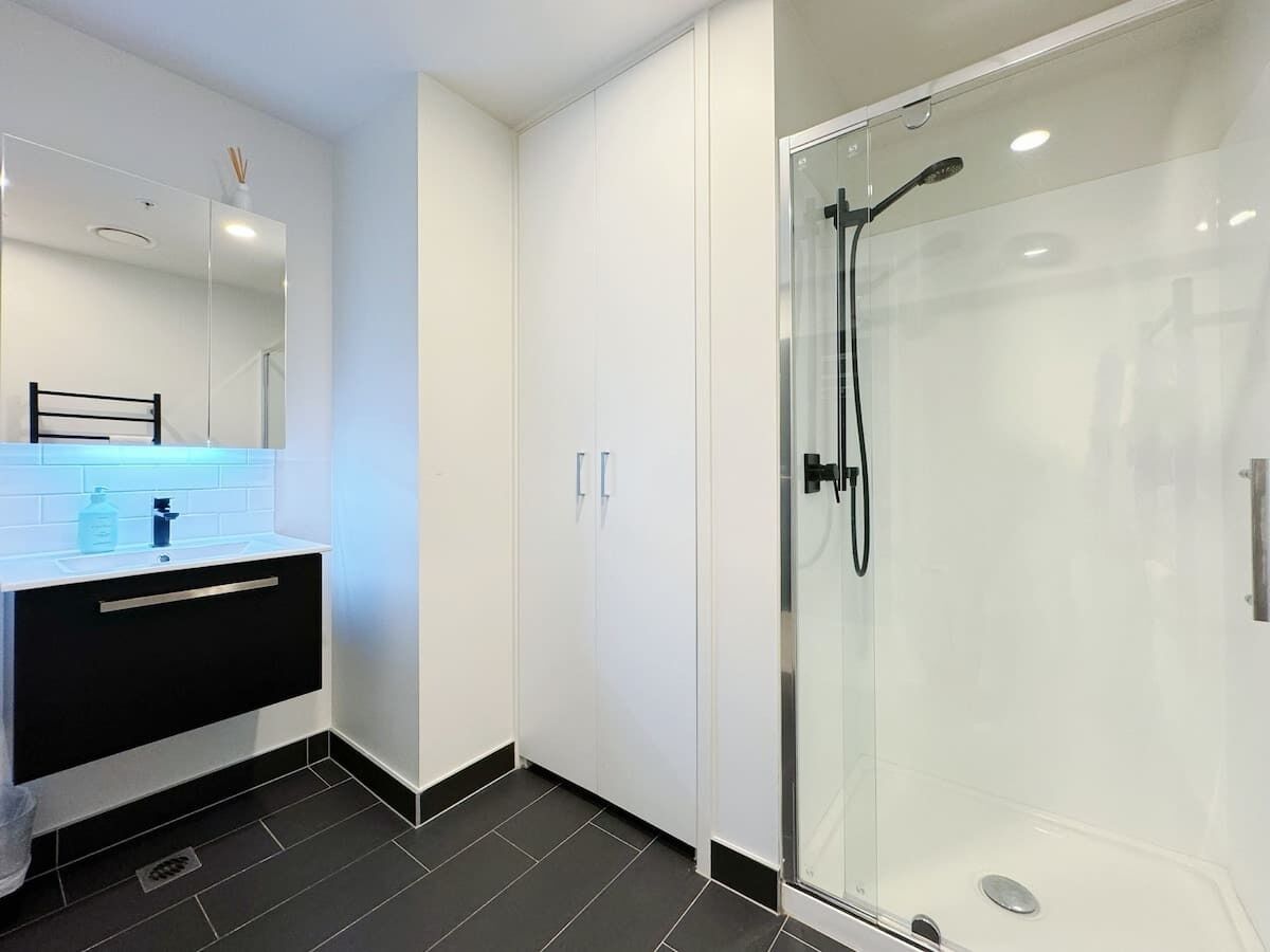Un baño moderno con ducha de vidrio, tocador blanco y lavamanos negro, además de un espejo sobre el tocador.