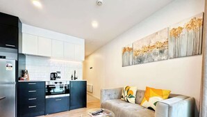 A modern living room with a grey sofa, a glass coffee table, and a kitchen area with white cabinets and a black refrigerator. - Central Location Cozy Studio with Pool Gym & Sauna Nearby Skytower (Auckland)