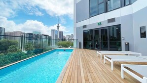 A rooftop pool with wooden deck and white lounge chairs, overlooking a cityscape with modern buildings and a tower. - Parkside Living Unique1B Apt in CBD vs Pool & Gym (Auckland)