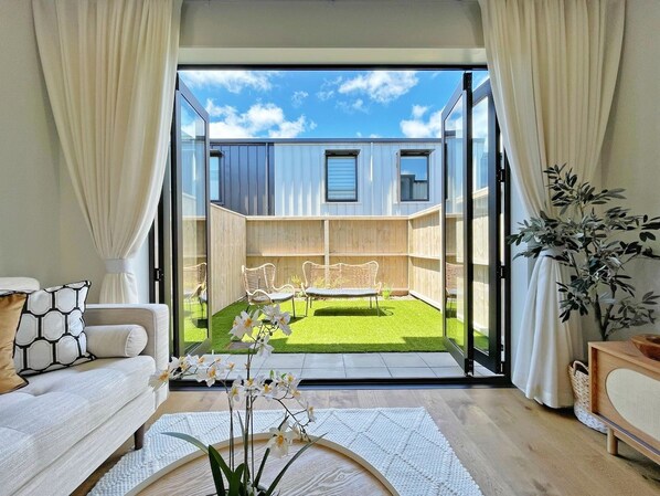 Una sala moderna con un gran ventanal corredera que da a un patio trasero con terraza de madera y dos sillones blancos de mimbre.