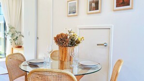 Ein Essbereich mit einem runden Glastisch, Korbstühlen und einer Vase mit getrockneten Blumen. An der Wand hängen gerahmte Bilder und im Hintergrund ist eine Tür zu sehen.