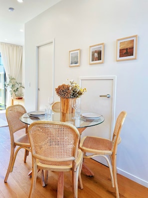 A dining area with a round glass table, wicker chairs, and a vase with dried flowers. There are framed pictures on the wall and a door in the background.