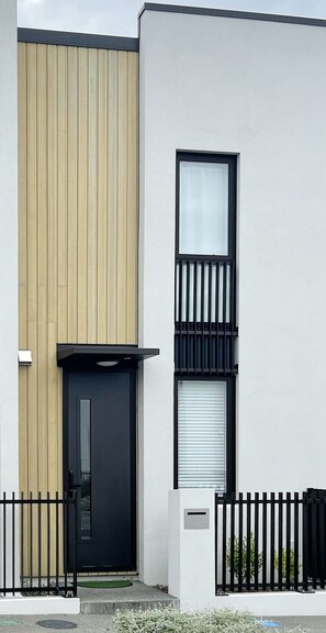 A modern two-story building with a black door, a vertical wooden panel, and a window with a metal grill.
