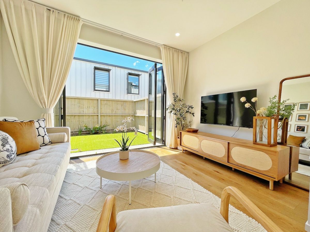 A modern living room with a sofa, coffee table, TV, and a view of a garden through large windows.