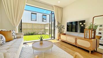 Una sala de estar moderna con sofá, mesa de centro, televisor y vista a un jardín a través de amplios ventanales.