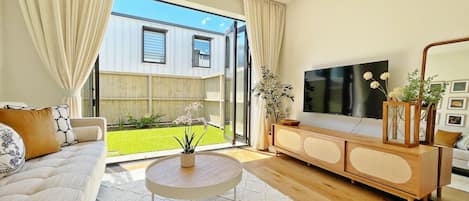 Una sala de estar moderna con sofá, mesa de centro, televisor y vista a un jardín a través de amplios ventanales.