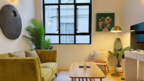 Ein modernes Wohnzimmer mit einem gelben Sofa, einem Holztisch, einem weißen Schreibtisch und einem großen Fenster mit schwarzen Rahmen.