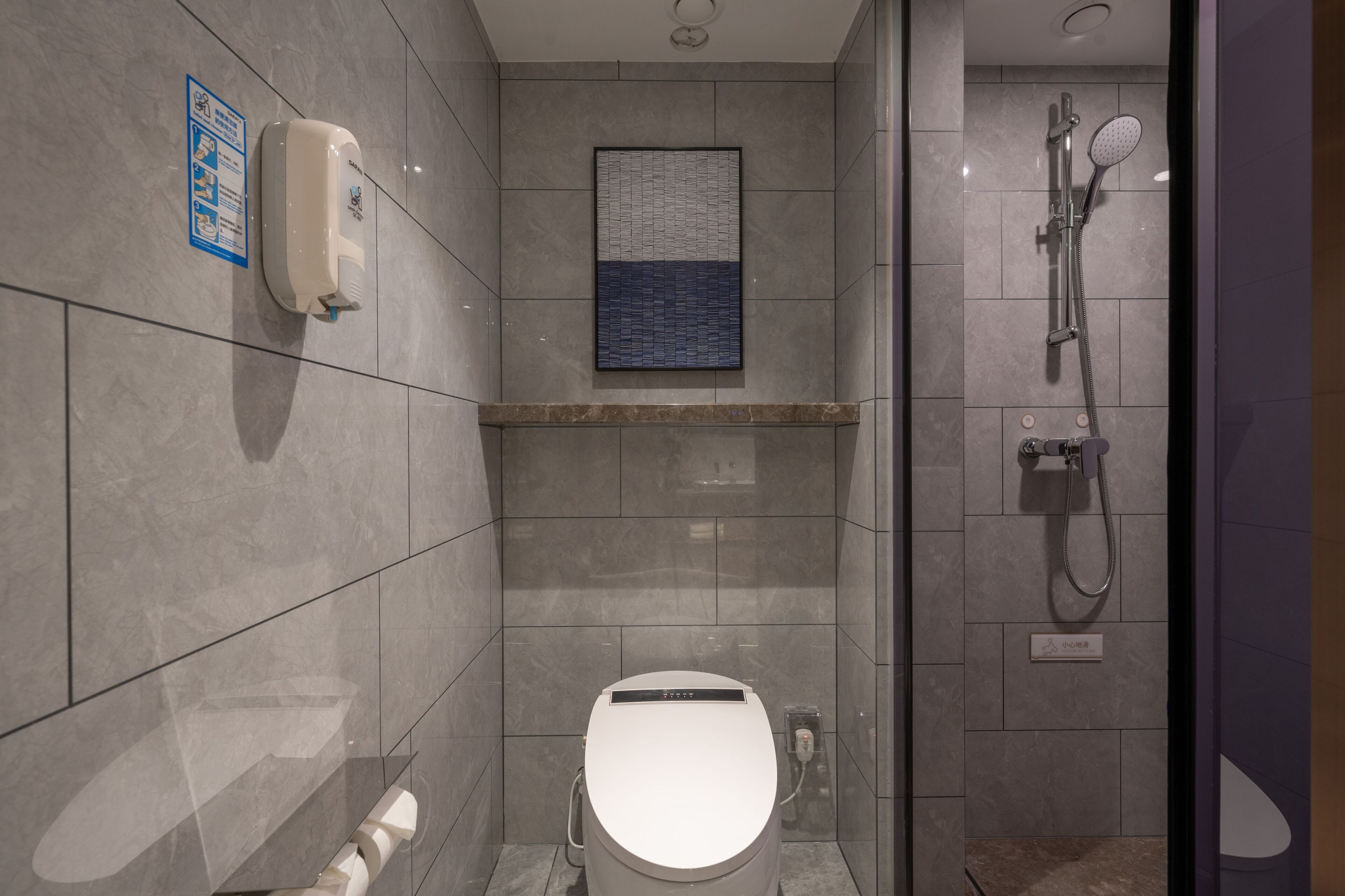A modern bathroom with a toilet, a hand dryer, a shower with a handheld showerhead, and a wall-mounted sign.