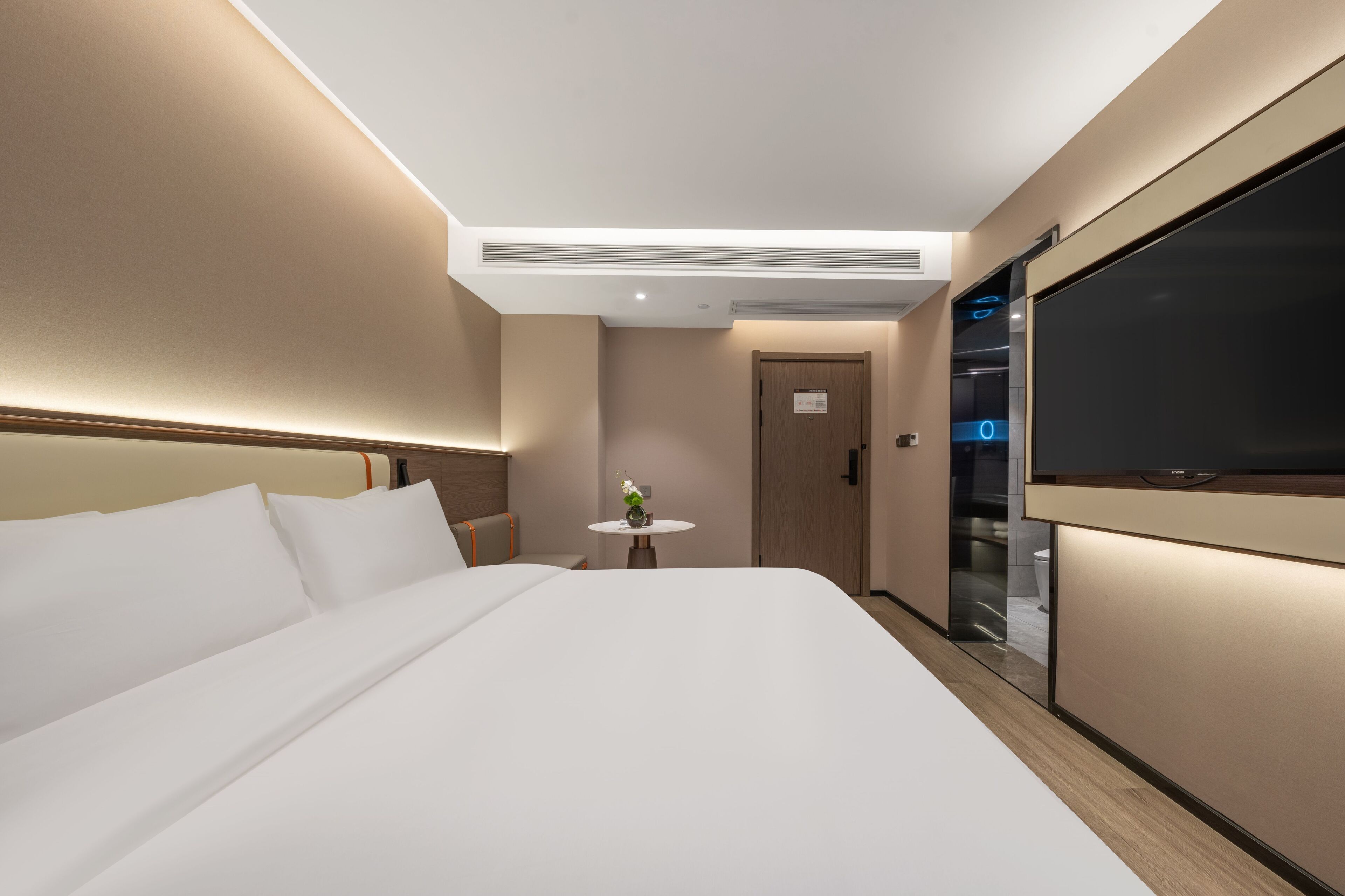 A modern hotel room with a large bed, a small round table with a vase, and a TV on the right wall.