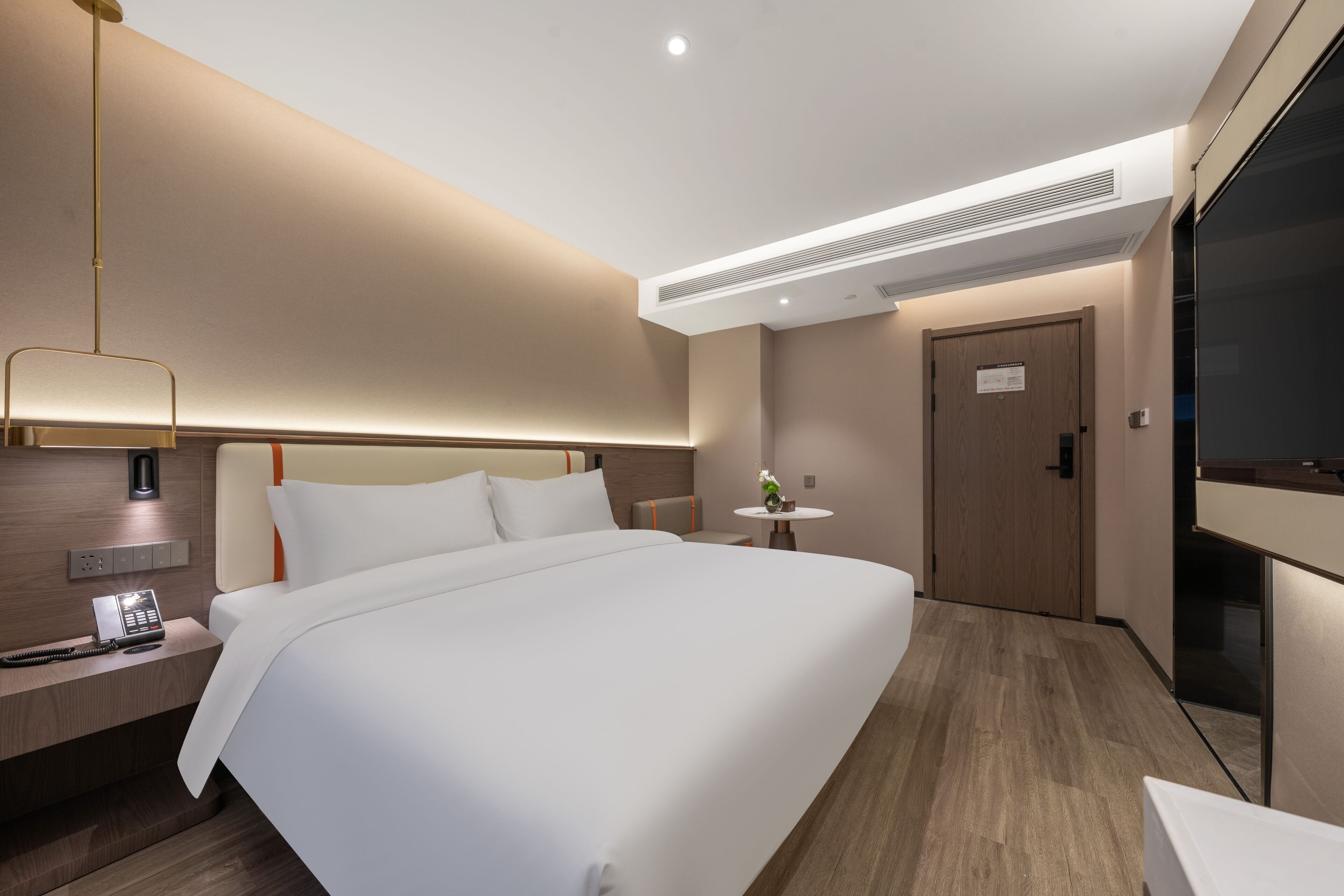 Ein modernes Hotelzimmer mit einem großen Bett, einem Nachttisch mit Telefon, einem kleinen runden Tisch mit Pflanze und einer Tür mit Schild.