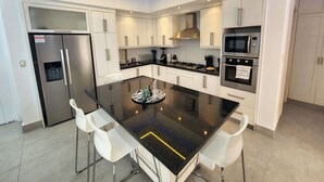 A modern kitchen with a central island, stainless steel appliances, white cabinetry, and bar stools. - Luxurious & Charming 3BR VILLA Palmas Del Mar Pool/Jacuzzi/BBQ in lovely Sosúa (Sosúa)