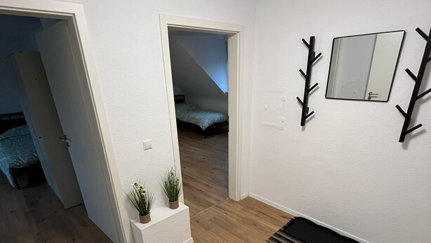 A modern, well-lit room with wooden flooring, a white wall, a small white planter with plants, a black coat rack, and a mirror. - nJoy! Business Apartment Pforzheim (Pforzheim)