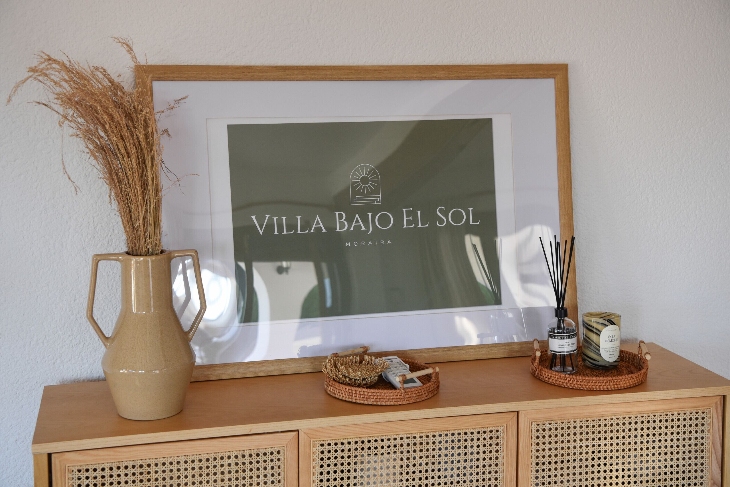 Ein gerahmtes Schild mit dem Schriftzug "Villa Bajo El Sol" und eine dekorative Vase mit getrockneten Pflanzen.