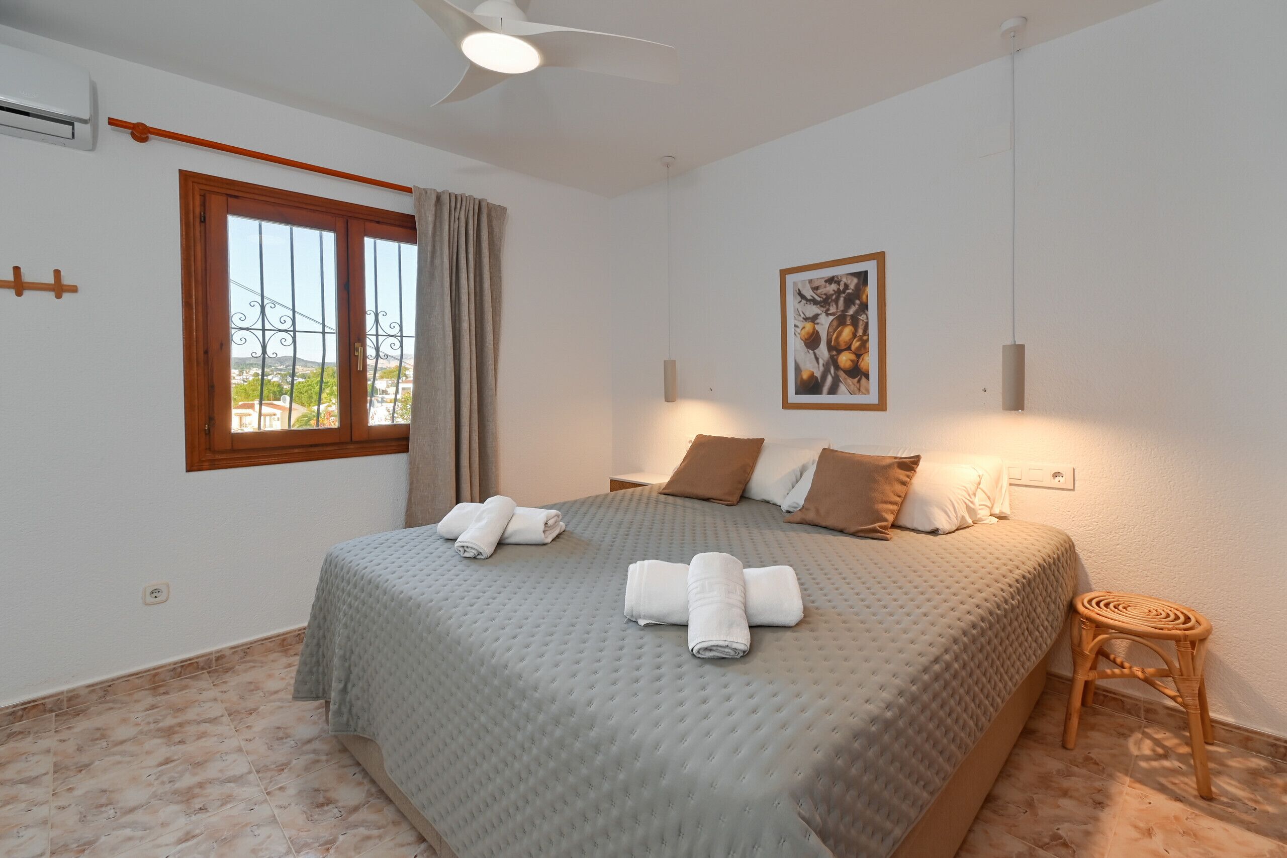 Un dormitorio con una cama grande, dos almohadas, un taburete pequeño, un cuadro en la pared y una ventana con cortinas.
