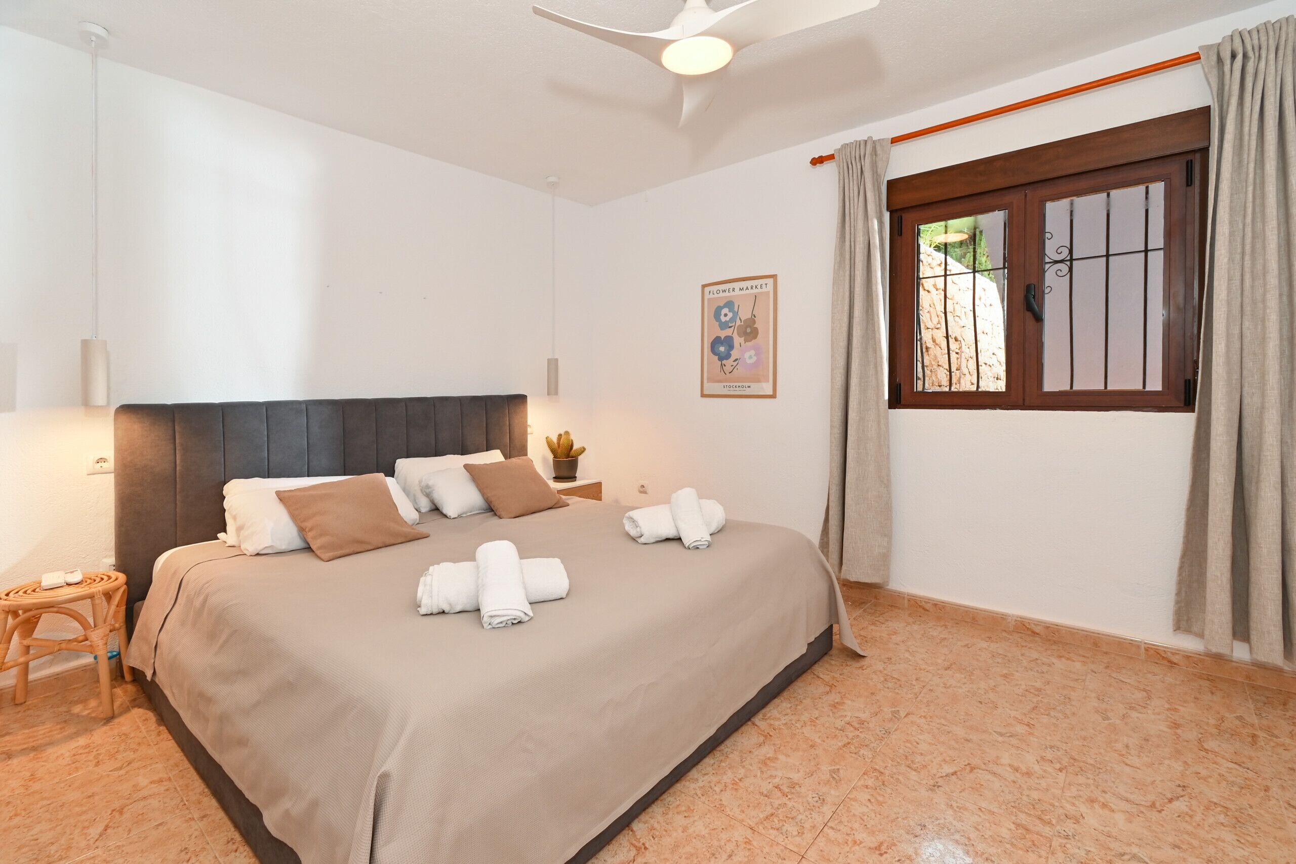 A neatly made bed with pillows and towels, a small wooden stool, a ceiling fan, and a window with curtains.