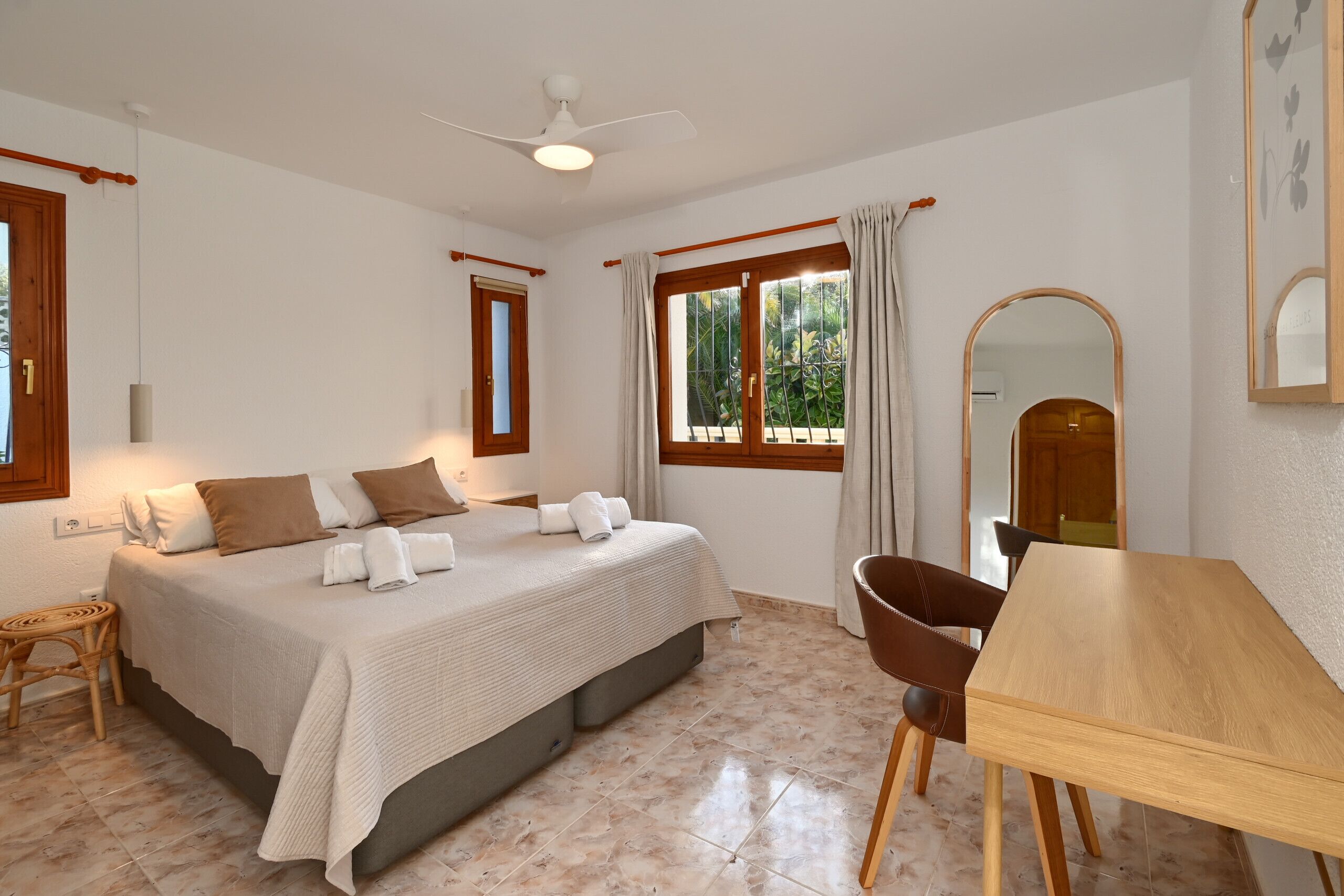 Une chambre à coucher avec un lit, une table en bois, une chaise, un ventilateur de plafond et une fenêtre avec des rideaux.